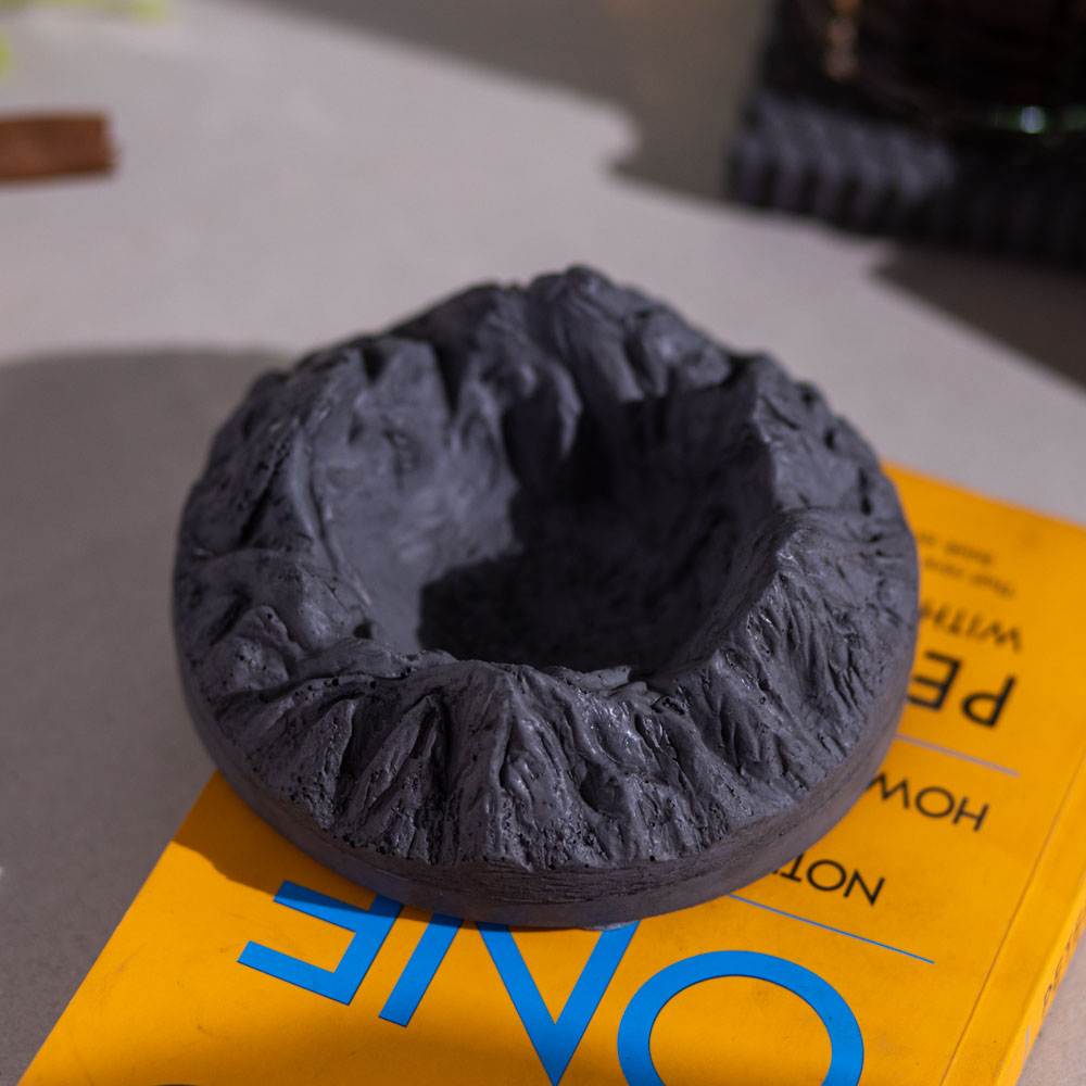 A round gray ashtray designed with mountainous patterns, placed on a yellow surface with a cigar resting on it.