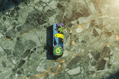 A small booklet of unbleached hemp rolling papers placed on a wooden surface, surrounded by green hemp leaves. The booklet features colorful artwork on the cover.