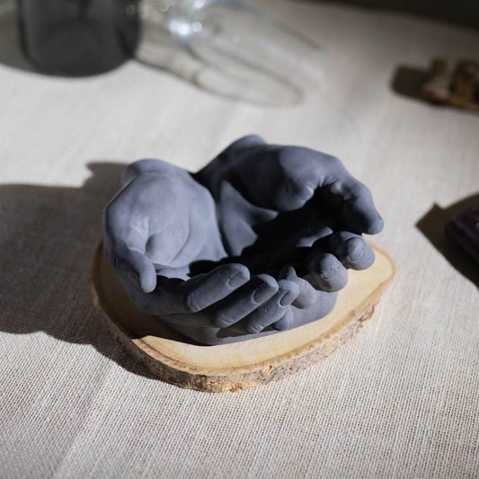 A hand-shaped, dark gray ceramic ashtray on a wooden base, placed on a beige fabric surface.