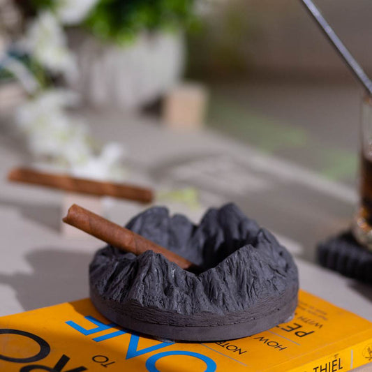A round gray ashtray designed with mountainous patterns, placed on a yellow surface with a cigar resting on it.