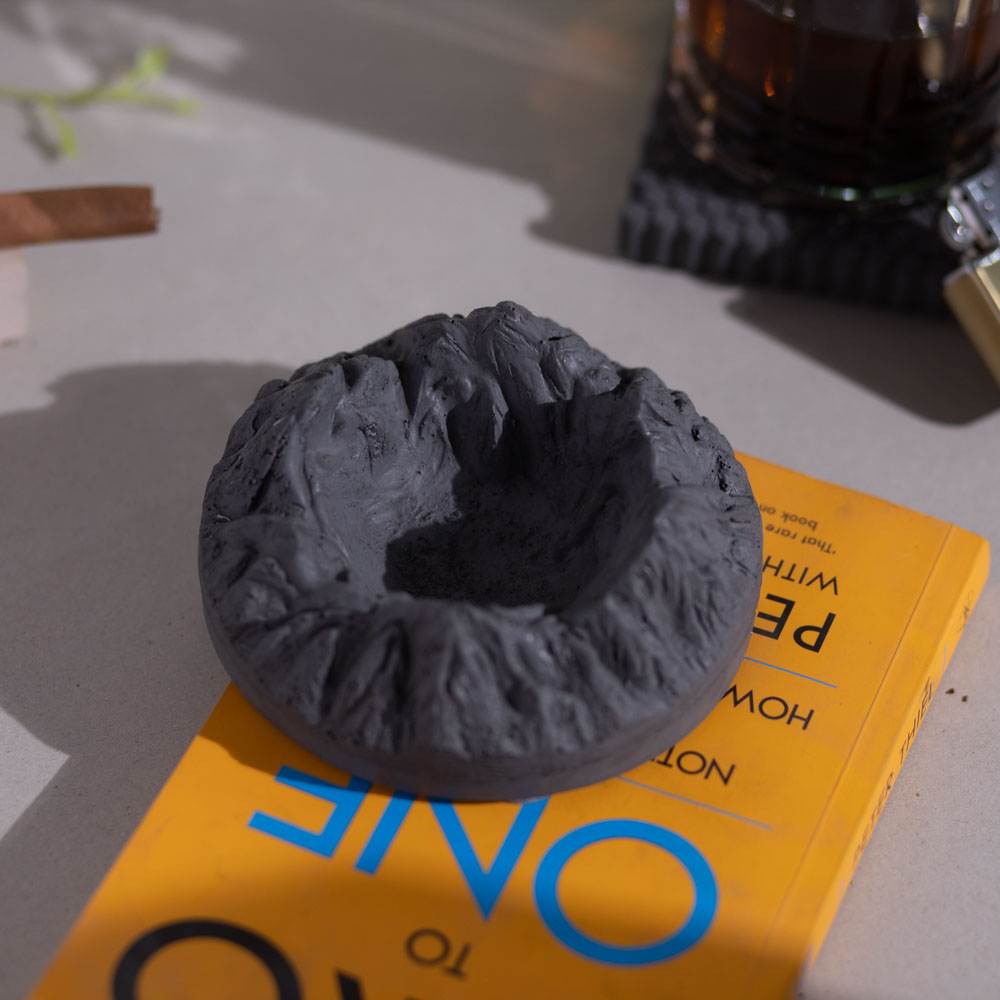 A round gray ashtray designed with mountainous patterns, placed on a yellow surface with a cigar resting on it.