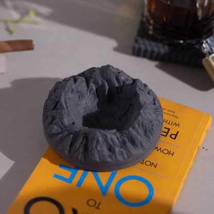 A round gray ashtray designed with mountainous patterns, placed on a yellow surface with a cigar resting on it.