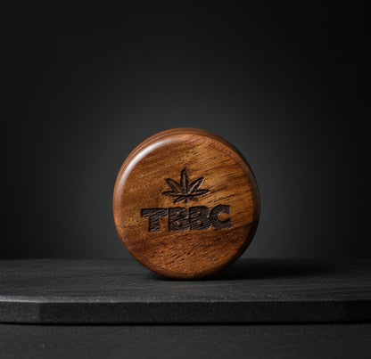 TBBC Signature 56mm wooden herb grinder closed to show the engraved lid and natural wood grain against a dark backdrop.