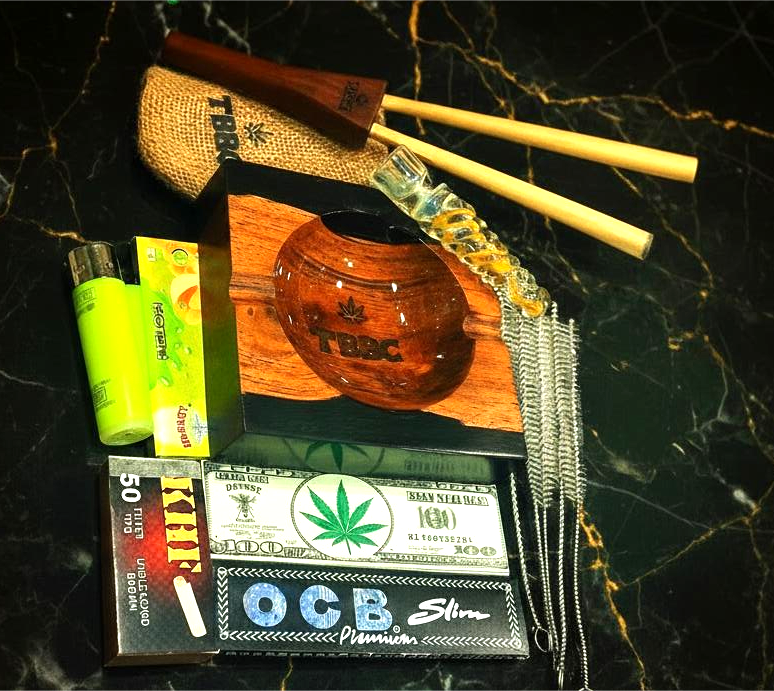 Collection of smoking accessories including pipes, glass chillum one hitter raw rolling papers,  KHF roachpads and lighters hornet 100 dollar paper, cleansing sticks on a dark marble surface with double barrel joint holder